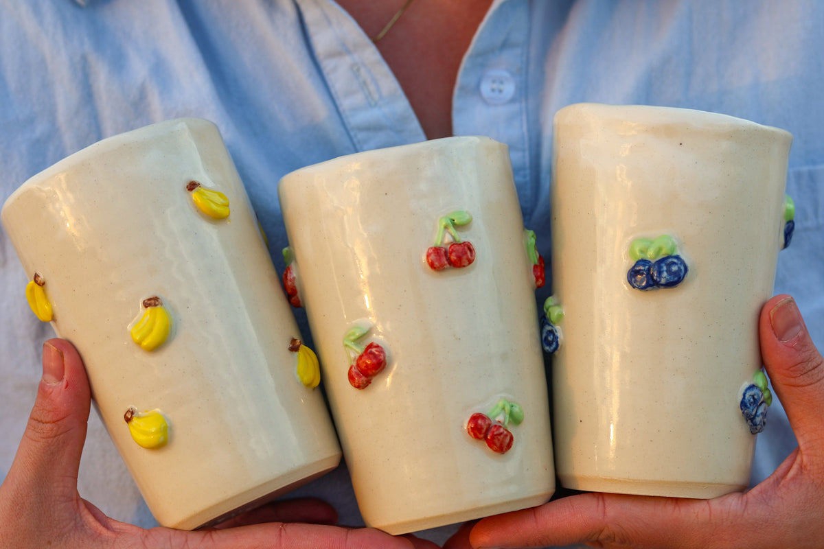 Three cream-colored ceramic tumblers decorated with colorful 3D fruit motifs (yellow bananas, red cherries, blue blueberries) held by hands wearing a light blue button-down shirt.