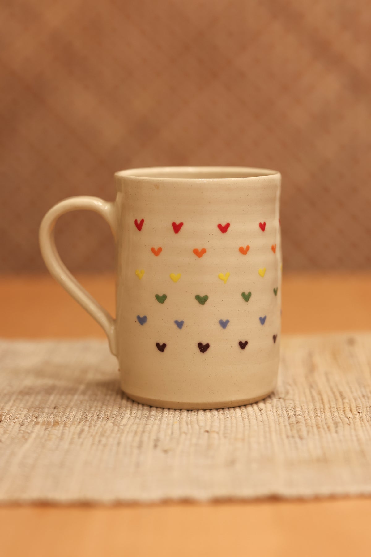 A beige ceramic mug decorated with rows of small hand-painted hearts in multiple colors, resting on a woven placemat against a warm brown textured background.