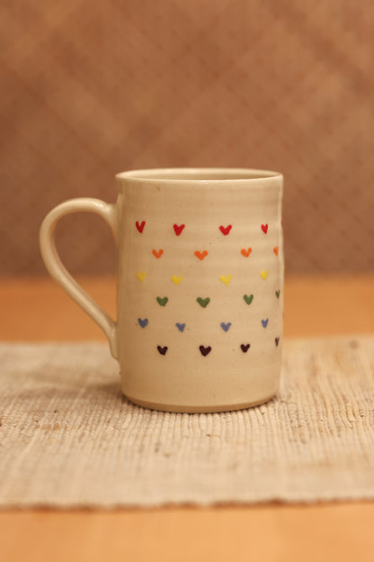 A beige ceramic mug decorated with rows of small hand-painted hearts in multiple colors, resting on a woven placemat against a warm brown textured background.