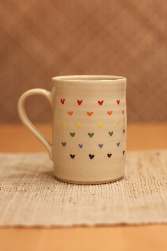 A beige ceramic mug decorated with rows of small hand-painted hearts in multiple colors, resting on a woven placemat against a warm brown textured background.