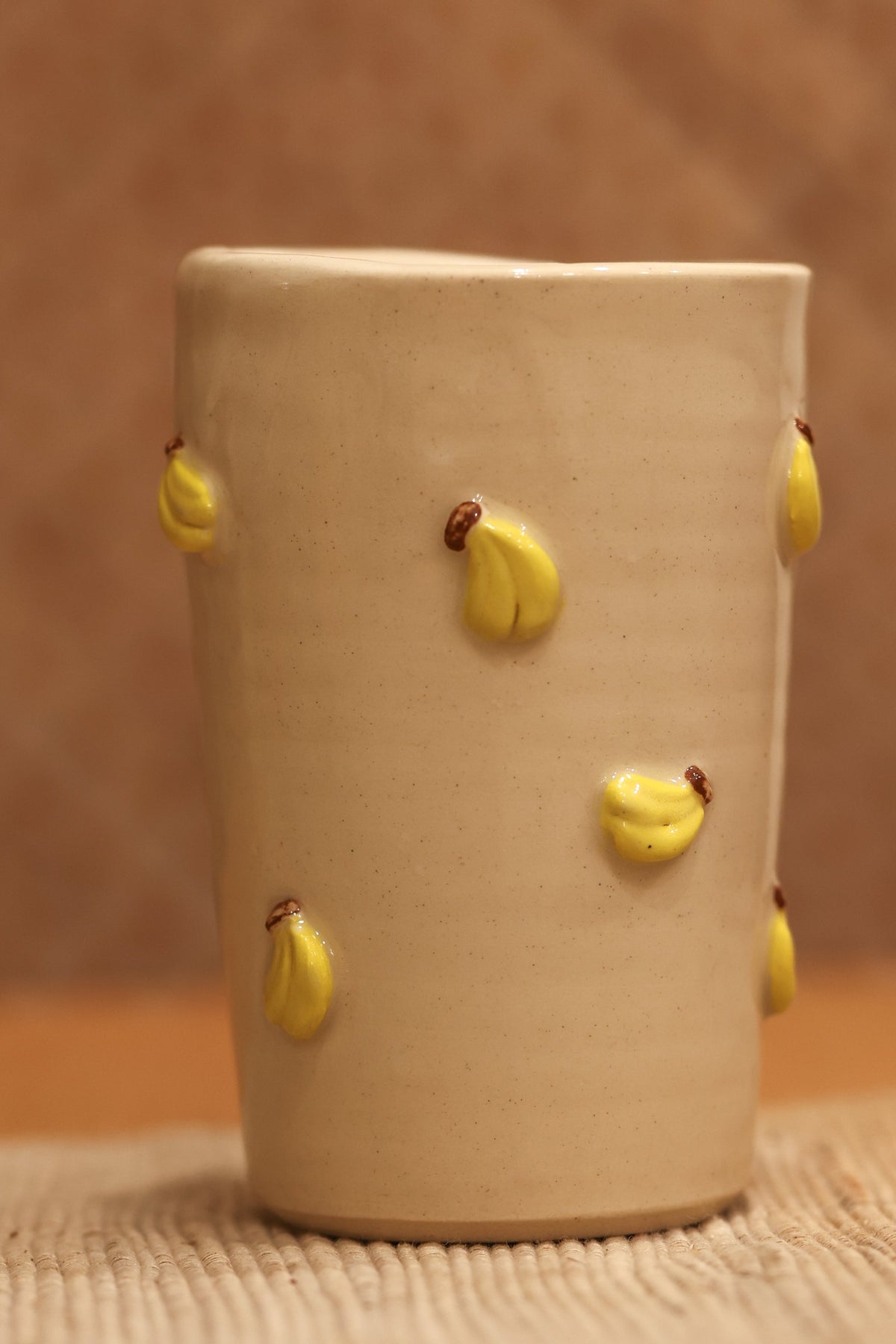 A handmade beige ceramic cup decorated with small yellow clay flower buds, set on a woven mat against a textured brown background.