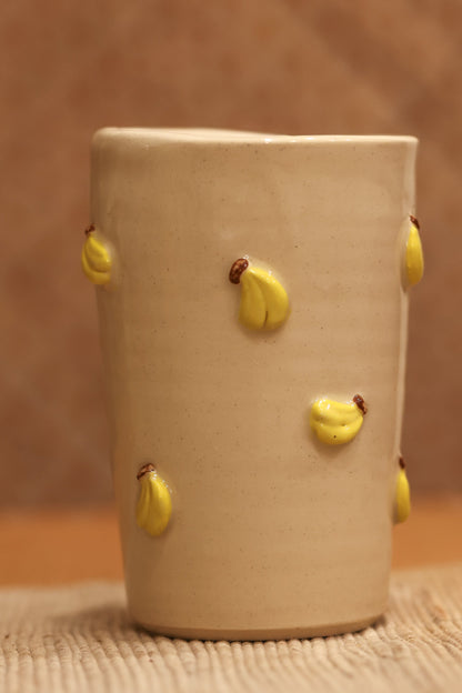 A handmade beige ceramic cup decorated with small yellow clay flower buds, set on a woven mat against a textured brown background.