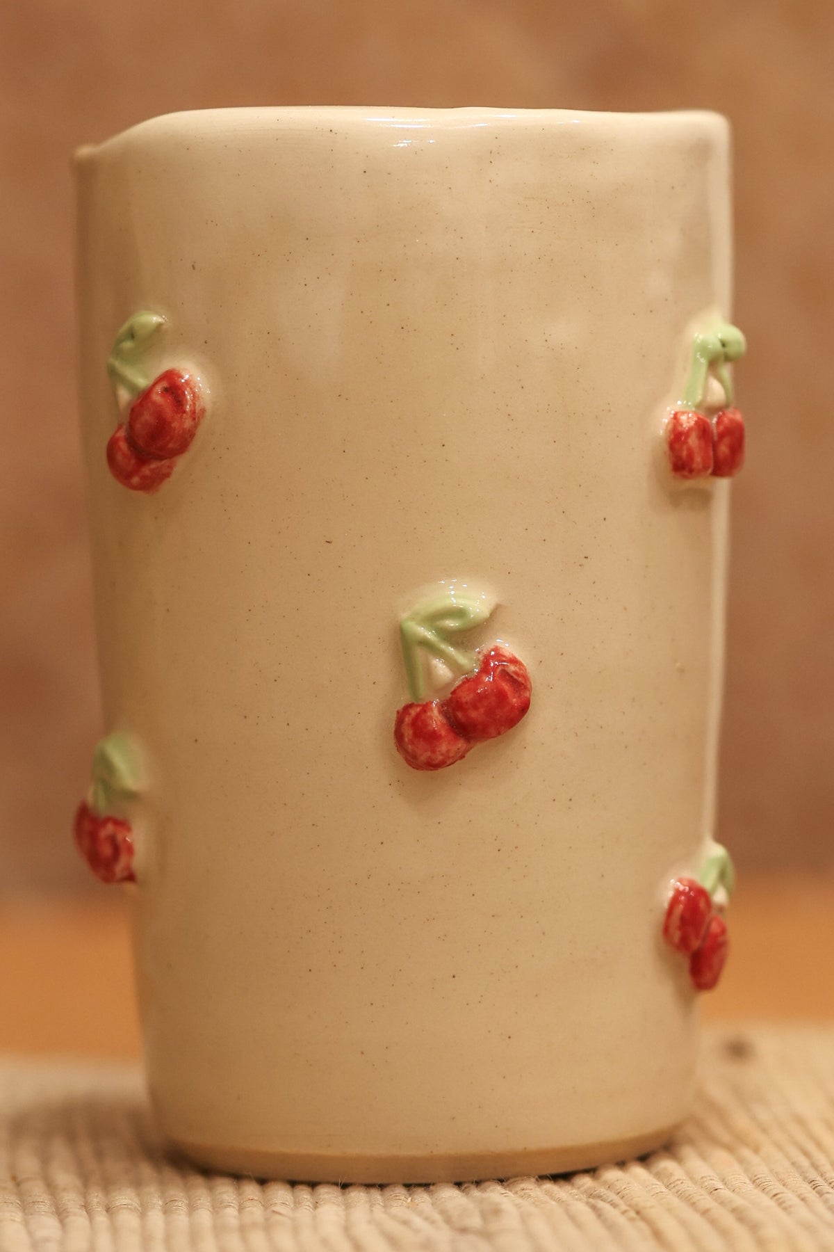 A small beige ceramic cup decorated with small red cherries with green leaves, set on a textured beige surface against a warm brown background.