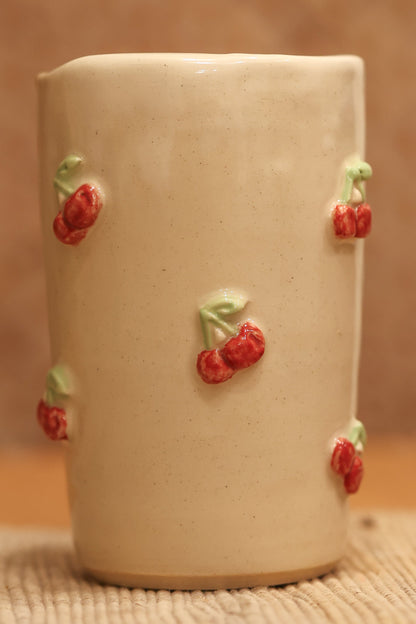 A small beige ceramic cup decorated with small red cherries with green leaves, set on a textured beige surface against a warm brown background.