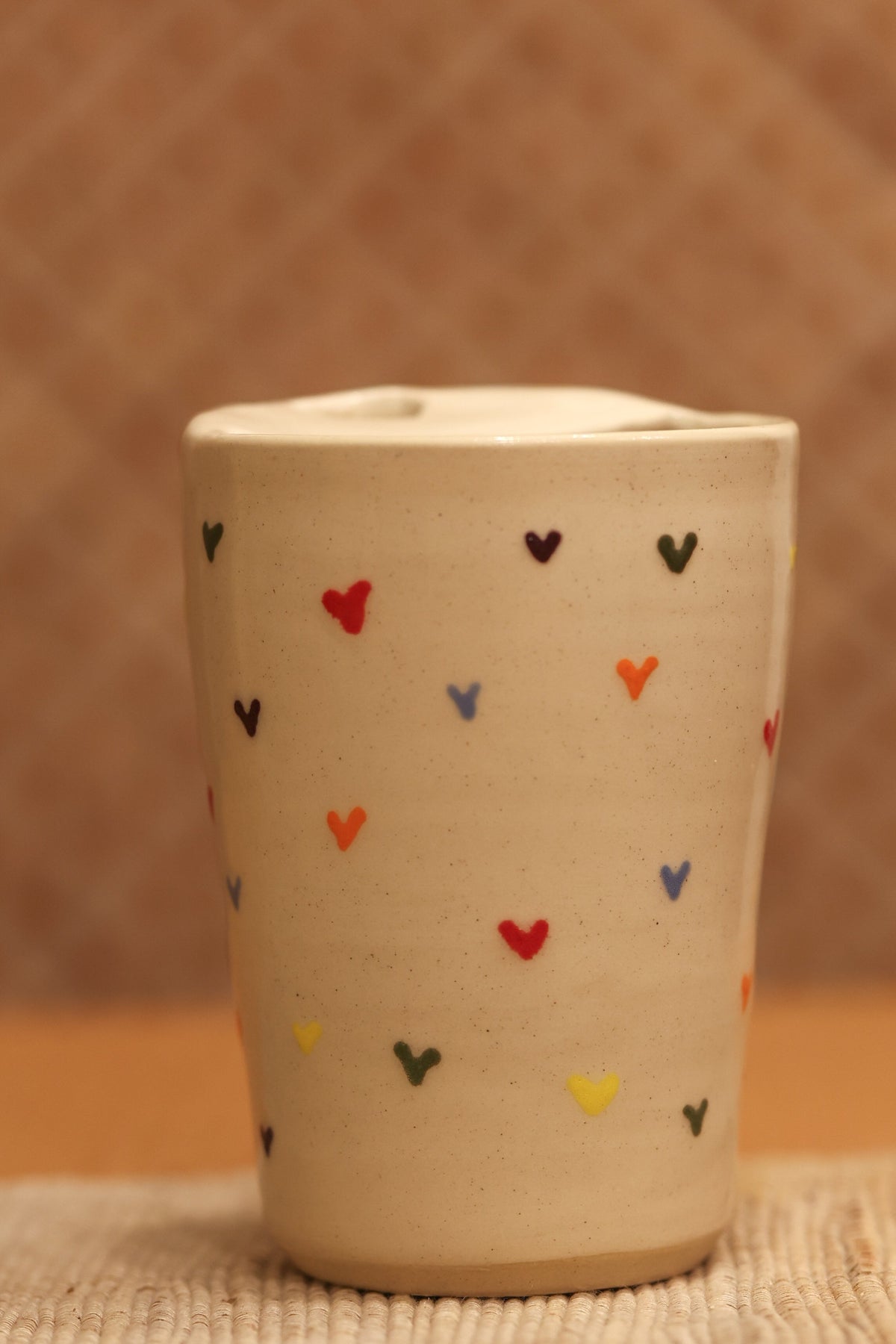 A small beige ceramic cup with a lid, decorated with small hand-painted hearts in red, blue, yellow, and black, set against a warm brown textured background.