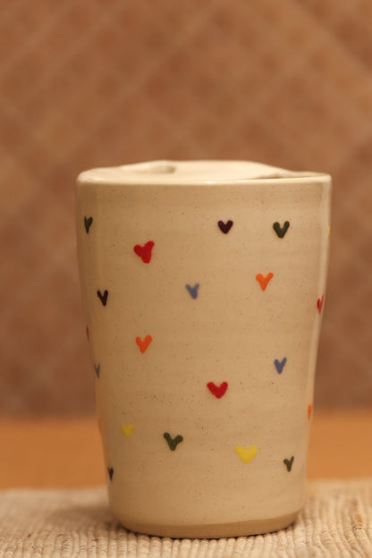 A small beige ceramic cup with a lid, decorated with small hand-painted hearts in red, blue, yellow, and black, set against a warm brown textured background.