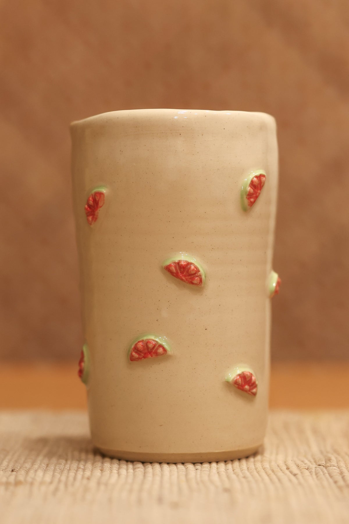 A small beige ceramic cup with small red and green strawberry motifs, standing on a woven mat against a warm brown textured background.