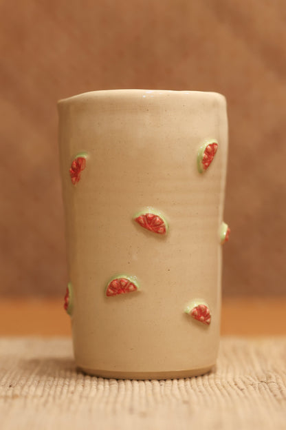 A small beige ceramic cup with small red and green strawberry motifs, standing on a woven mat against a warm brown textured background.